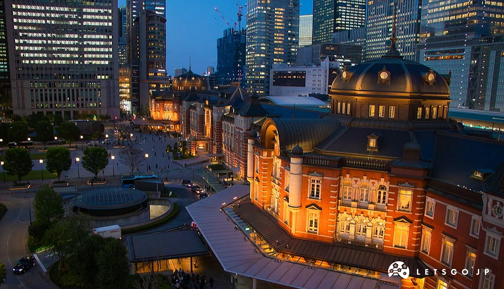 東京車站紅磚建築夜景,建築與後方現代高樓群皆亮起暖黃燈光。